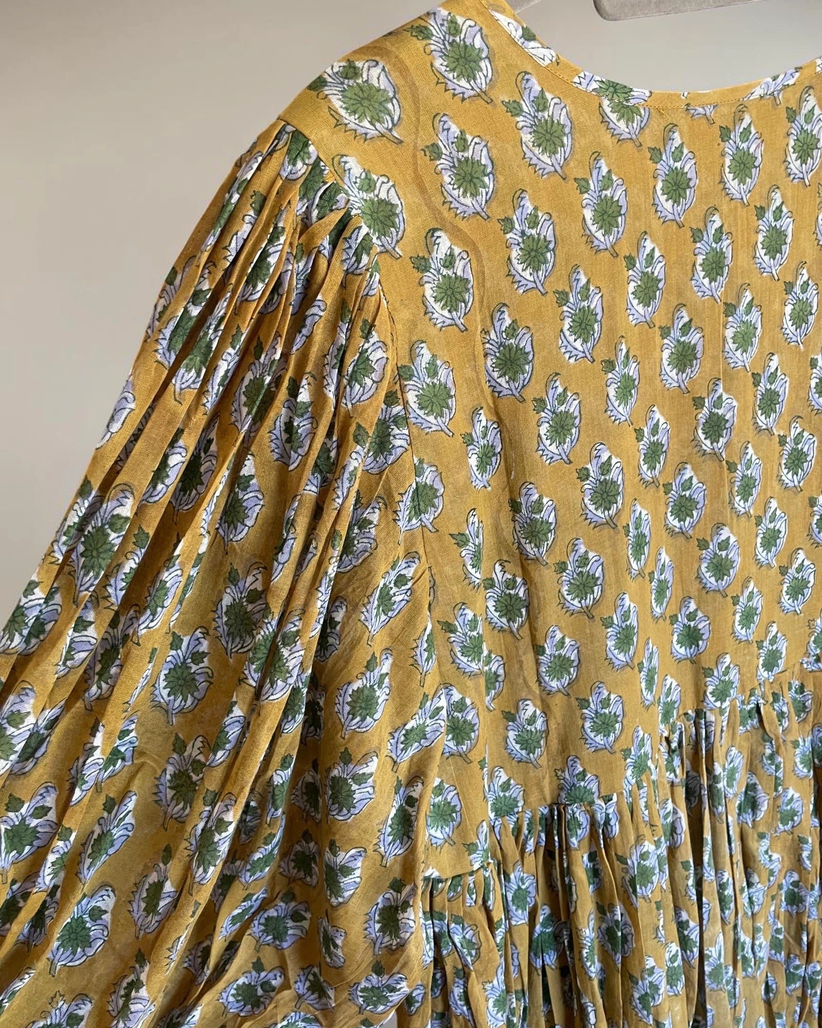 A close-up of a mustard yellow dress with a green and white paisley block print pattern, featuring pleated long sleeves and a gathered waist.