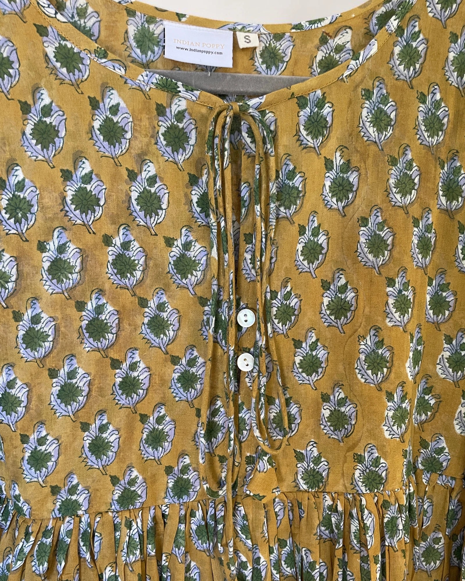 A close-up of a mustard yellow blouse with a green and white leaf block print, featuring a tie-neck detail and mother of pearl, white buttons.