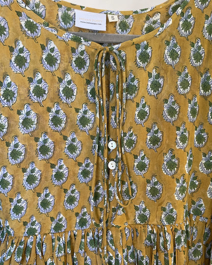 A close-up of a mustard yellow blouse with a green and white leaf block print, featuring a tie-neck detail and mother of pearl, white buttons.