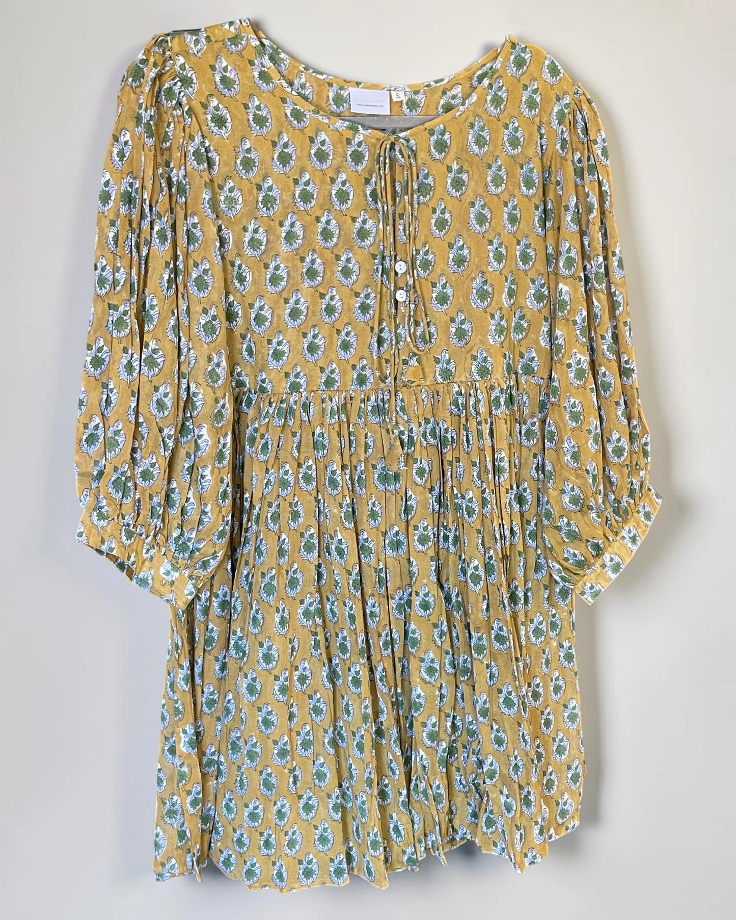 A yellow short-sleeve tunic dress with a green and blue floral pattern, featuring a tie-neck detail and a gathered waist.