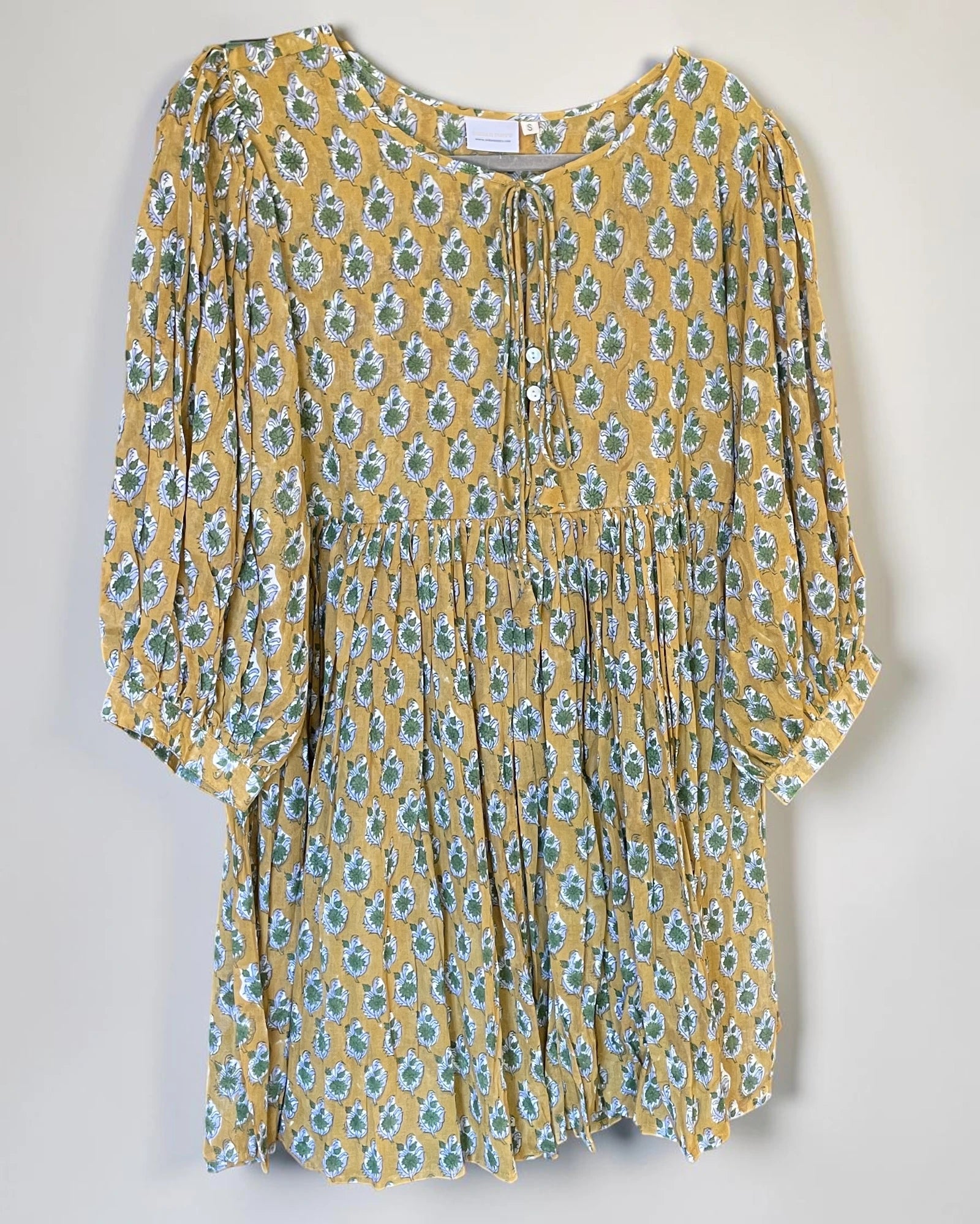A yellow short-sleeve tunic dress with a green and blue floral pattern, featuring a tie-neck detail and a gathered waist.
